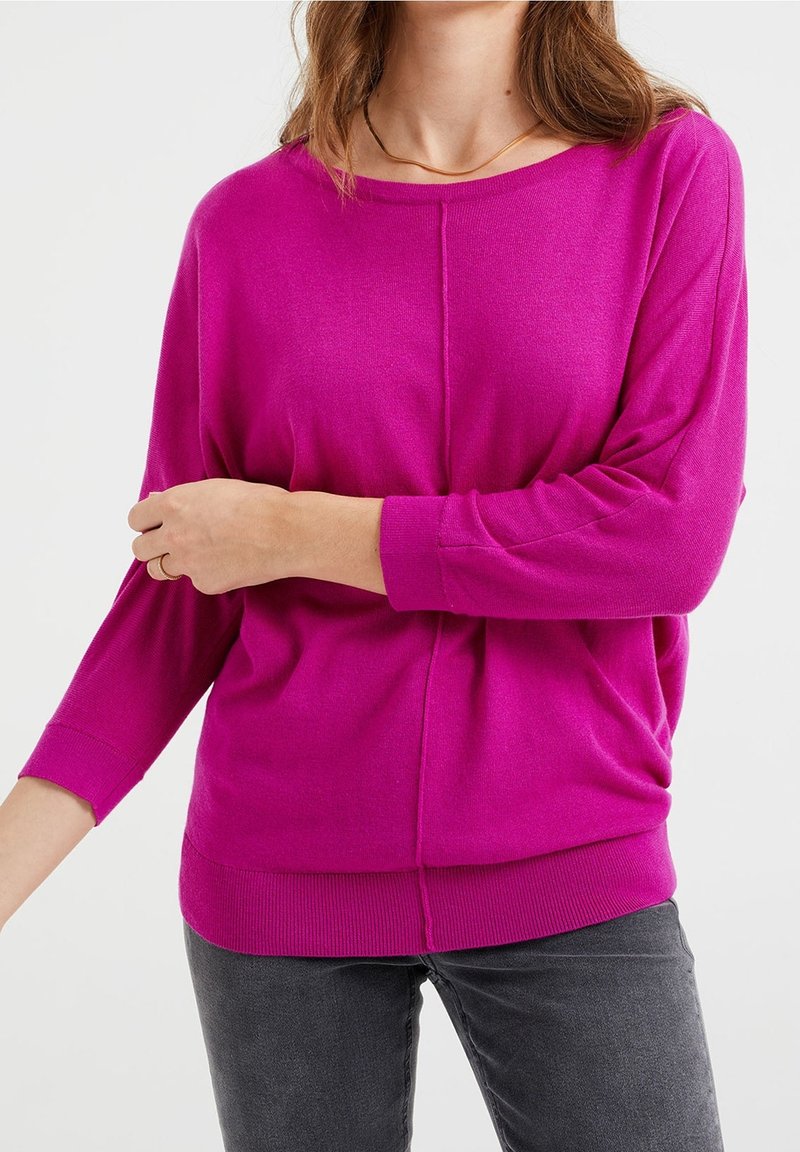 Femme portant un haut à manches longues rose vif avec poignets et ourlet côtelés, et un pantalon gris foncé, debout devant un fond clair.