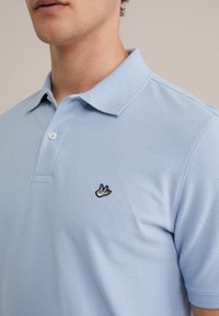 Gaiši zils polo krekls, izgatavots no teksturēta auduma. Iekļauj klasisku apkakli, trīs pogu aizdari un melnu izšuvumu ar logotipu uz krūtīm.