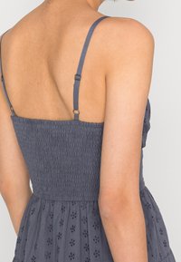 Šedé šaty se smyčkovaným živůtkem, tenkými ramínky a vyšitými detaily. Rozšířená sukně s výřezem, který ukazuje texturu a design.