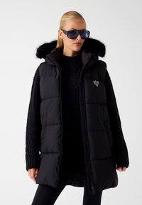 Juodas pūkinis liemenė su pūkiniu gaubtu. Turi siūtos dalis, per didelį siluetą ir raukšlėtus megztus rankovių galus. Uždarymas užtrauktuku su logotipo paiste.