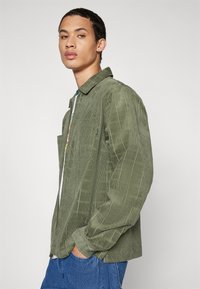 Grøn kordfløjlsskjorte med ternet mønster, der har krave, forsigtig lomme og lynlås foran. Matcher med blå jeans.