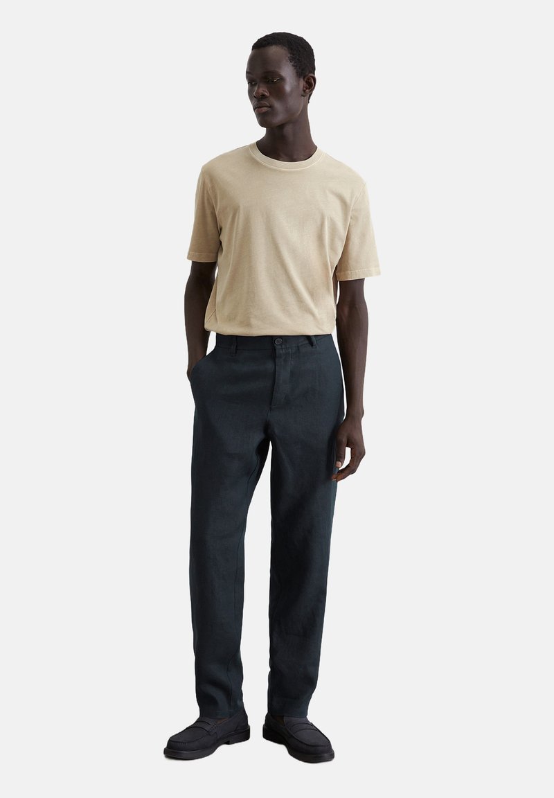 Beige katoenen t-shirt met korte mouwen, gecombineerd met donkergrijze linnen broek. De outfit wordt aangevuld met donkere instappers.