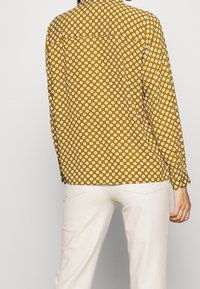 Camicia a maniche lunghe color senape con motivo floreale bianco, caratterizzata da un colletto button-down e un orlo curvo. Abbinata a pantaloni beige.