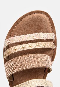 Zbliżenie brązowego sandała z czterema dekoracyjnymi paskami: różowym tweedem, złotymi ćwiekami, r&oacute;żowo-złotymi rhinestonami i kremowym materiałem teksturowanym.