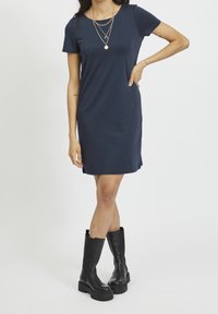 Granatowa sukienka z krótkim rękawem, wykonana z gładkiej tkaniny, z okrągłym dekoltem i luźnym krojem, noszona z czarnymi, wysokimi na kolana, masywnymi botkami.