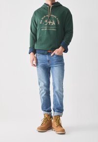 Sudadera verde con gráfico beige, camisa de cuadros azul por debajo, jeans azul claro y botas de trabajo color beige con cordones amarillos.