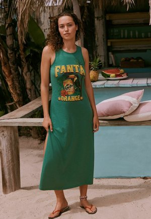 Kobieta w zielonej sukience bez rękawów z logo Fanta Orange, stoi boso na piaszczystym podłożu obok drewnianego stołu z owocami i poduszkami.
