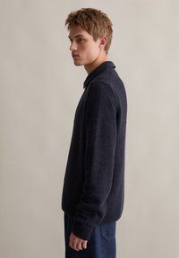 Ciemnoniebieski sweter o texturedze z kołnierzykiem, prążkowanymi mankietami i dołem. Posiada długie rękawy i luźny krój, zestawiony z dżinsowymi spodniami.