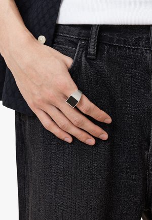 Silver metal ring with a geometric design, featuring a rectangular black stone inlay. Worn on a hand against dark denim fabric.