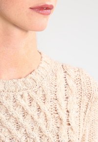 Beige gestrickter Pullover mit einem Zopfmuster, der über einen Rundhalsausschnitt und eine weiche, strukturierte Oberfläche verfügt. Nahaufnahme des Halsbereichs.