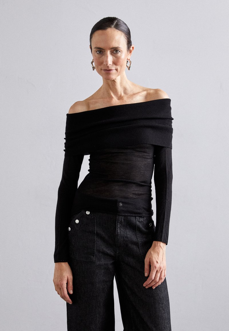 Melna off-shoulder blūze ar slāņotu ribu adījumu; apvienota ar augstās jostas līnijas tumšām džinsu biksēm ar sudraba pogām.