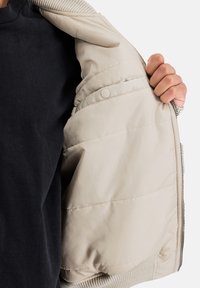 Interior de una chaqueta acolchada color beige claro con puños acanalados, que presenta una textura suave y lisa y un cierre de botón oculto en el forro interior.