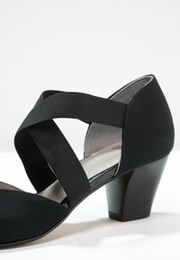 Chaussure noire à talon avec un bout rond, dotée de sangles élastiques croisées et d'un dessus en tissu texturé. Le talon est solide et de forme bloquée.