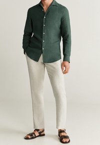 Camisa de manga larga verde oscuro con textura suave y cuello, combinada con pantalones beige claros y sandalias negras de tiras.