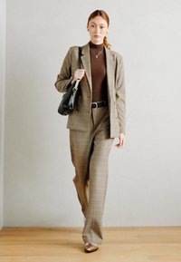 Costume à carreaux marron avec une veste ajustée et un pantalon à jambes larges, associé à un pull col roulé marron et des chaussures dorées. Sac à main noir comme accent.