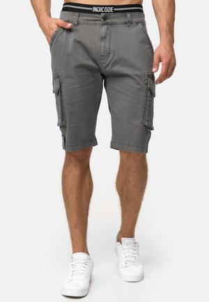 Man draagt grijze cargoshorts met zijzakken, een zwarte tailleband met het label "INDICODE," en witte sneakers, staand tegen een effen achtergrond.