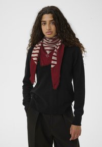 Kvinde med krøllet hår iført en sort sweater og mørke bukser, accessoriseret med et rødt og beige stribet tørklæde mod en ensfarvet hvid baggrund.