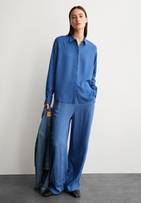 Blauwe satijnen longsleeve shirt gecombineerd met bijpassende wijde broek. Geaccentueerd met een casual gedragen denimjacket. Textuurstof met een gladde afwerking.