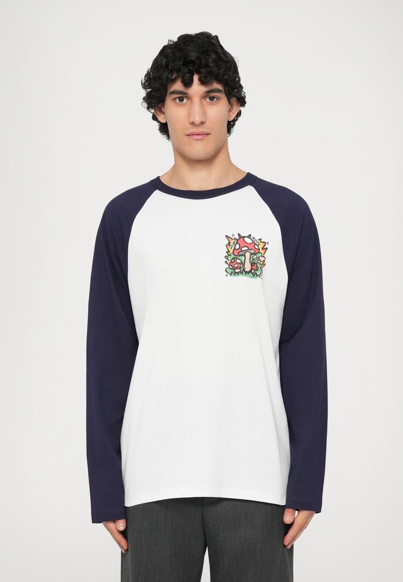 T-shirt à manches longues en raglan avec un corps blanc et des manches bleu marine, affichant un graphique de champignon coloré sur la poitrine. Matière en coton, coupe décontractée.