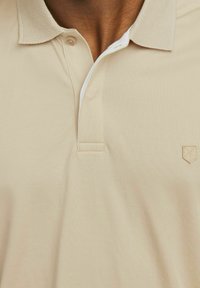 Beige polo krekls ar tekstūrētu apkakli, diviem pogām un izšūtu logo uz krūšu. Gluda auduma un brīvas pieguves.