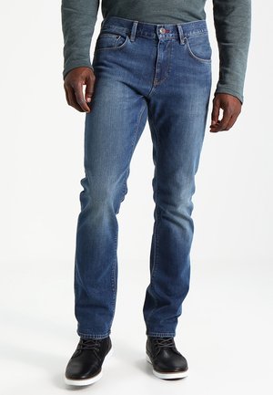 Mann trägt blaue Jeans, graues Langarmshirt und schwarze Schuhe und steht vor einem einfarbigen weißen Hintergrund.