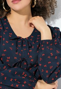 Marineblaue Bluse mit roten Schleifenmustern, ausgestattet mit einem V-Ausschnitt, Kordelzugdetails und ballonförmigen Ärmeln mit elastischen Bündchen. Glatte Textur.