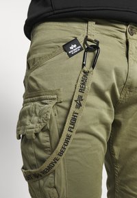 Pantalonii cargo de culoare verde măsliniu au o etichetă neagră cu marcă, un clips tip carabină și o curea decorativă "Remove Before Flight" cu text negru.