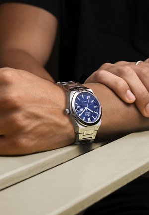 Strieborné nerezové hodinky s modrým ciferníkom, striebornými hodinovými značkami a dátumovou funkciou na prepojenej náramku, umiestnené na zápästí.