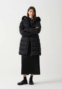 Zwarte donsjas met een afneembare bontvoering aan de capuchon, ritssluiting en geribbelde boorden, gedragen over een lange zwarte rok en schoenen.