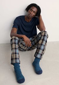 Mornarska majica, karirane hlače z bež, modro in črno barvo ter modre nogavice s temnimi konicami, sedeče na svetli tla.