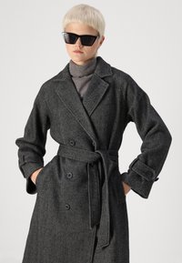 Abrigo largo de pata de gallo en blanco y negro, con cinturón, botones grandes y solapas anchas. La modelo lleva un cuello alto gris y gafas de sol.