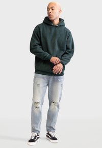 Felpa verde scuro con tasca a marsupio e coulisse regolabile, abbinata a jeans chiari strappati e sneakers bianco e nere.