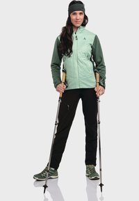 Hellgrüne und dunkelgrüne Jacke, schwarze Hosen, hält Trekkingstöcke. Strukturiertes, tailliertes Design; knielang mit Taschen. Grüne Wanderschuhe.