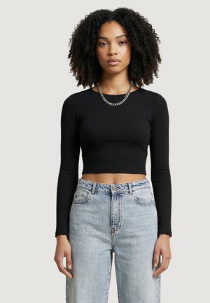 Giovane donna con capelli ricci naturali che indossa un top nero a maniche lunghe corto, una collana a catena argento e jeans azzurri a vita alta.