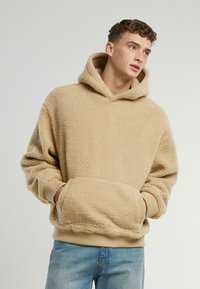 Beige fleece hettegenser med teksturert overflate, stor frontlomme og ribbestrikkede mansjetter. Har en justeerbar hette og en avslappet passform.