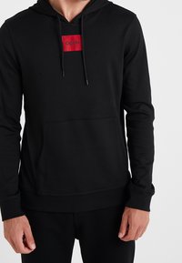 Sudadera con capucha negra, hecha de una tela suave, que cuenta con un bolsillo frontal, puños acanalados y una etiqueta roja con "HUGO" en el pecho.