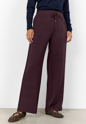 Frau trägt dunkelviolette, weit geschnittene Hose mit Kordelzug, schwarzes Oberteil und braune Slip-On-Schuhe mit Nieten, stehend auf grauem Boden.