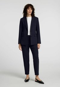 Marineblå skreddersydd dress med en blazer med hakket revers, to frontlommer og matchende smale bukser; kombinert med en hvit topp og svarte leiligheter.