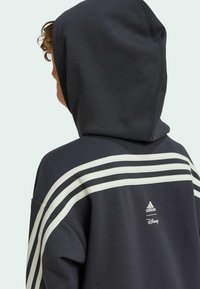 Felpa grigia scura con un ampio cappuccio, design a tre strisce bianche sulla schiena e loghi di Adidas e Disney sul retro in basso.