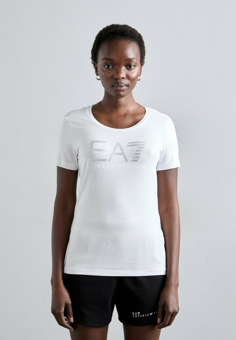 T-shirt bianca a maniche corte con scollo rotondo, decorata con il testo argentato "EA7" e "EMPORIO ARMANI" impreziosito da piccoli strass.