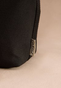 Svart texturerat tyg med en liten beige tygetikett med texten "Cath Kidston London" på kanten, placerad mot en beige yta.