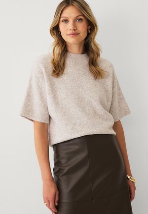 Kvinde iført en lys beige kortærmet sweater og en højtaljet mørkebrun lædernederdel står mod en ensfarvet baggrund.