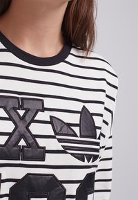 Žena oblečená v bílém a černém pruhovaném topu s velkými černými písmeny s koženou texturou a logem Adidas trefoil na hrudi.