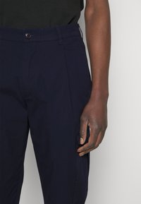 Marina blå byxor i ett lättviktigt material med knappstängning, sidoficka och strukturerad design. Slät textur och normalt snitt.