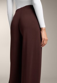 Brązowe spodnie szerokie, wykonane z gładkiego, elastycznego materiału. Posiadają wysoki stan i subtelne detale szwów z tyłu. Połączone z dopasowaną białą bluzką.