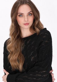 Schwarzer Strickpullover mit strukturiertem, offenem Maschenmuster, langen Ärmeln und lockerer Passform. Das Modell posiert mit verschränkten Armen.