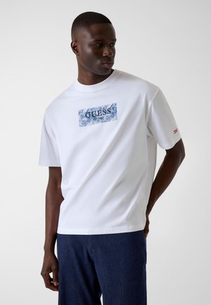 Mann trägt weißes Guess T-Shirt mit blauem Logoaufdruck und dunkelblaue Hose, schaut nach links mit einer Hand hinter dem Rücken.