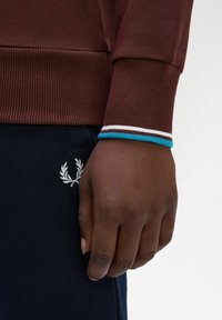 Felpa bordeaux con polsini a coste, con righe color teal, bianco e marrone; pantaloni blu navy con logo ricamato bianco; mano di carnagione scura appoggiata.