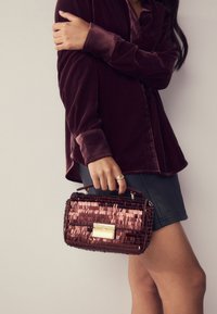 Handtasche aus Paillettenmaterial mit rechteckiger Form. Sie verfügt über einen goldenen Verschluss und einen oberen Griff. Kombiniert mit einer burgunderroten Samtbluse.