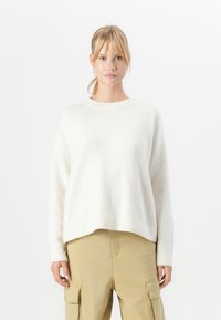 Giovane donna con capelli biondi raccolti in una coda di cavallo, che indossa un maglione bianco a maniche lunghe e pantaloni cargo beige, in piedi davanti a uno sfondo bianco semplice.